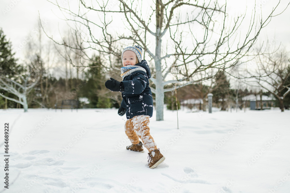 Adorable toddler boy having fun in a backyard on snowy winter day. Cute child wearing warm clothes playing in a snow.