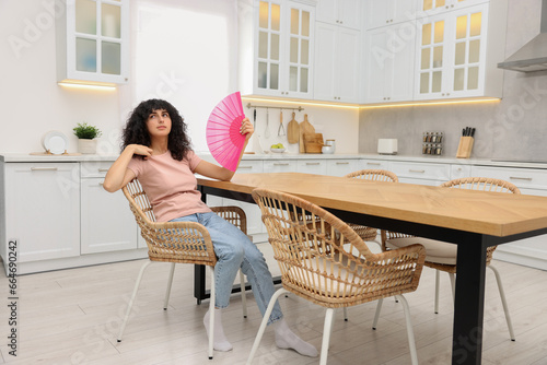 Young woman waving pink hand fan to cool herself at table in kitchen