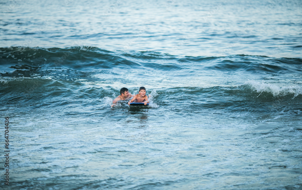 Little surfer learn to ride on surfboard on sea wave. Father with son play in summer ocean, learning surfing. Little boy swim on  surf board