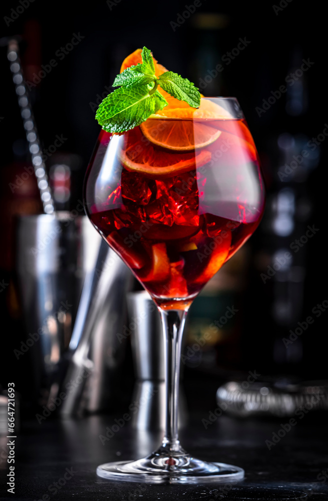 Red sangria cocktail drink with tempranillo wine, peach, plum, orange, lime and ice. Black bar counter background, steel bar tools and bottles