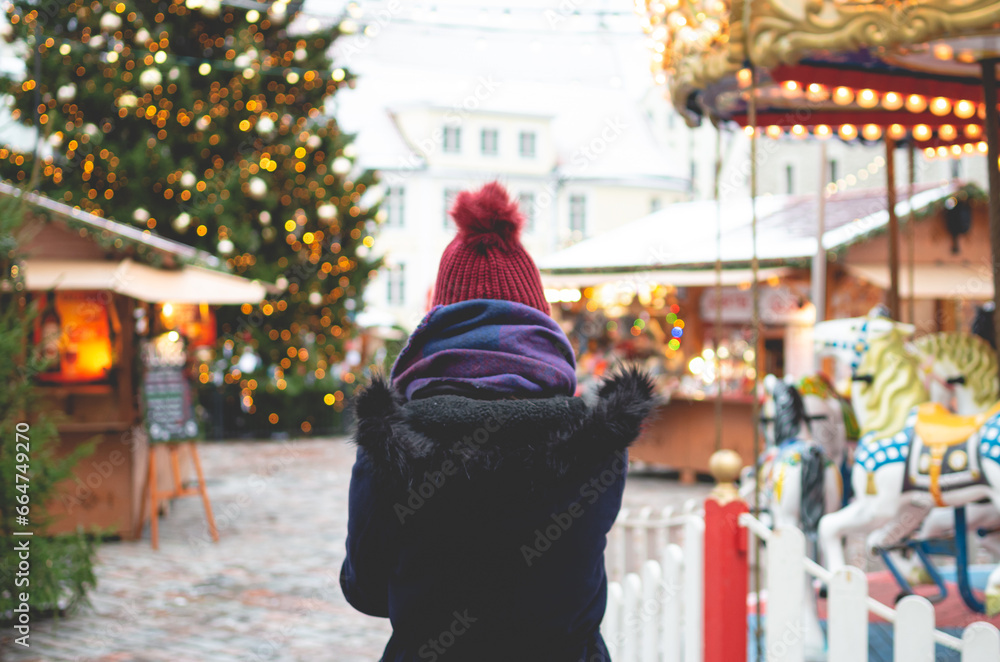 A young woman in a blue winter coat and a red hat standing on a Tallinn Town Hall Square on a cold winter day with a Christmas tree and a Christmas fare in the background