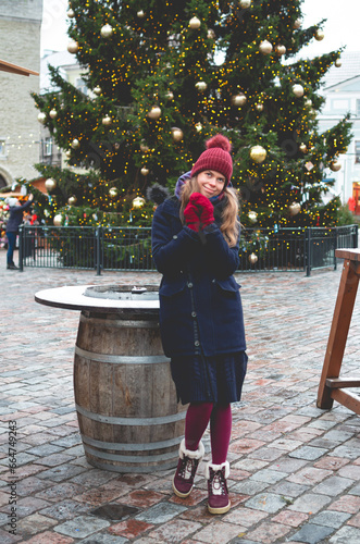A young handsome smiling woman dreaming while holding hands together and standing on a Tallinn Town Hall Square on a cold winter day with a Christmas tree in the background