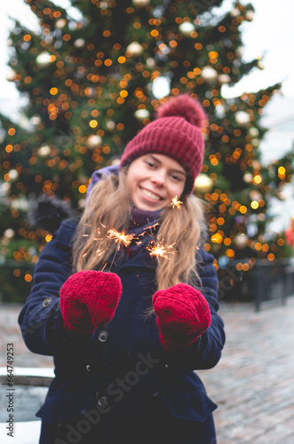 A young handsome smiling woman holding sparklers and standing on a Tallinn Town Hall Square on a cold winter day with a Christmas tree in the background