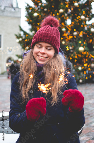 A young handsome smiling woman holding sparklers and standing on a Tallinn Town Hall Square on a cold winter day with a Christmas tree in the background