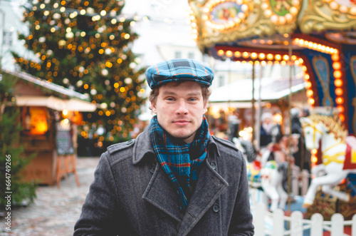 A handsome young man in a grey coat with a Scottish tweed cap and tartan scarf standing on a Tallinn Town Hall Square with a Christmas tree and a Christmas fare in the background