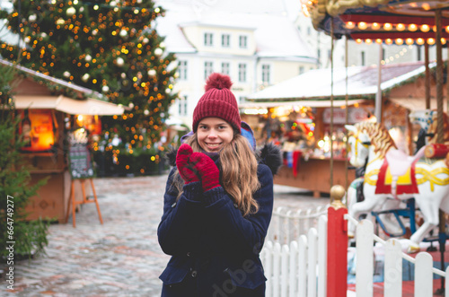 A handsome young woman in a blue winter coat, red hat and gloves holding hands together and standing on a Tallinn Town Hall Square with a Christmas tree and a Christmas fare in the background