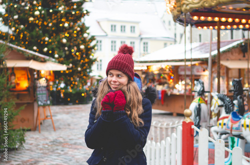 A handsome young woman in a blue winter coat, red hat and gloves holding hands together and standing on a Tallinn Town Hall Square with a Christmas tree and a Christmas fare in the background