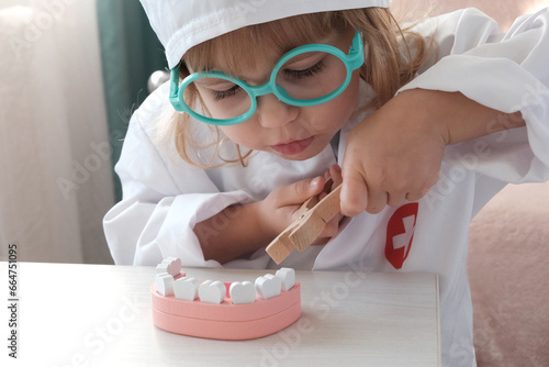 Children's game of dentist. A little European girl in a doctor's costume and glasses holding a toy in her hands pulls out a tooth on a jaw model. Dental oral hygiene concept.