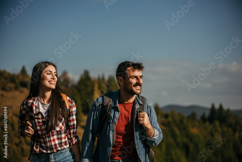 A diverse couple with radiant smiles embarking on an adventurous hike, carrying vibrant backpacks on a sun-drenched day