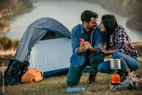 Diverse couple camping by a lake with a stove, capturing the moment on a phone