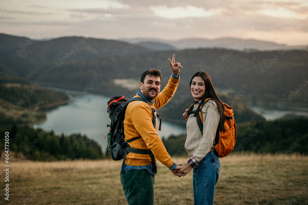 Smiling embraced hikers with backpacks taking a walk on a hill, admiring a faraway lake