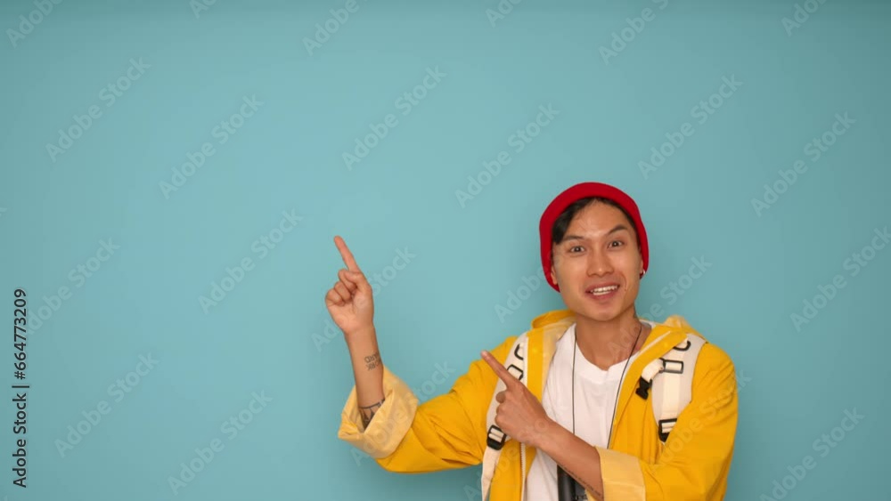 Portrait of a young Asian man in a yellow jacket and red cap pointing with his finger to the copy space on a blue background Portrait of a young Asian man in a yellow jacket and red cap pointing with his finger to the copy space on a blue background