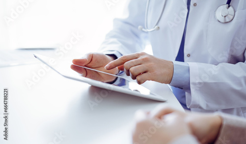 Doctor and patient sitting at the table in clinic. The focus is on female physician s hands using tablet computer  close up. Medicine and healthcare concept