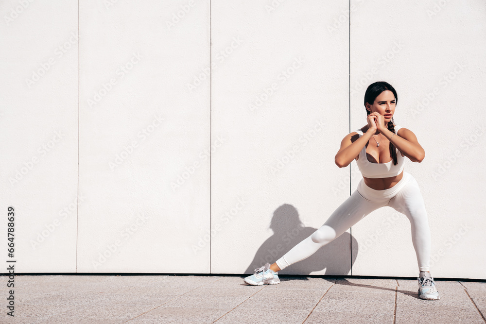 Fototapeta premium Fitness woman in sports clothing. Sexy young beautiful model athlete doing fitness workout. Female making exercises in the street at sunny day. Stretching out before training. Makes lunges near wall