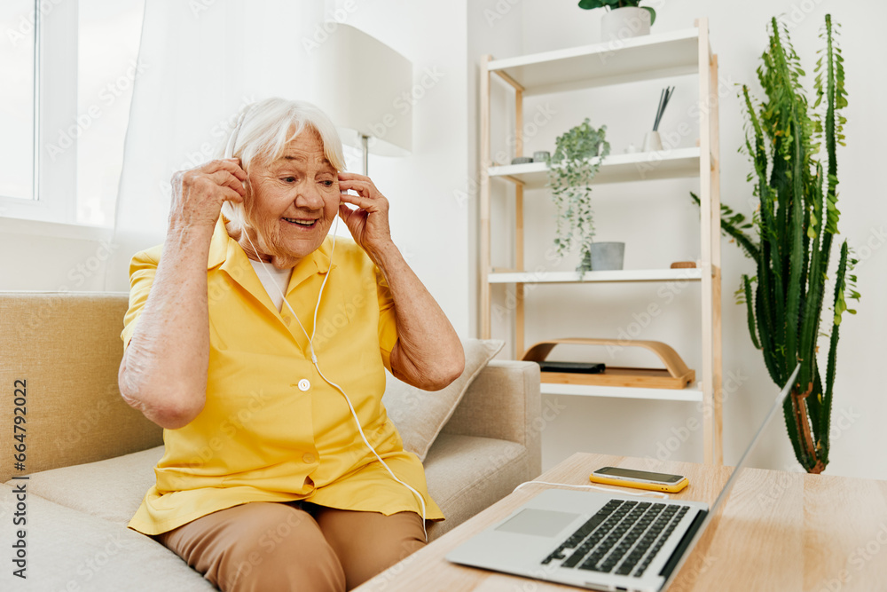 Elderly woman wearing headphones with a laptop communicating online by video call smile, sitting on the couch at home and working in a yellow shirt, the lifestyle of a retired woman. Elderly woman wearing headphones with a laptop communicating online by video call smile, sitting on the couch at home and working in a yellow shirt, the lifestyle of a retired woman.