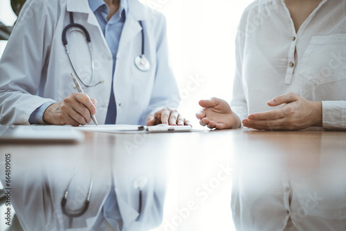 Doctor and patient sitting near each other at the wooden desk in clinic. Female physician's filling up a records form. Medicine concept
