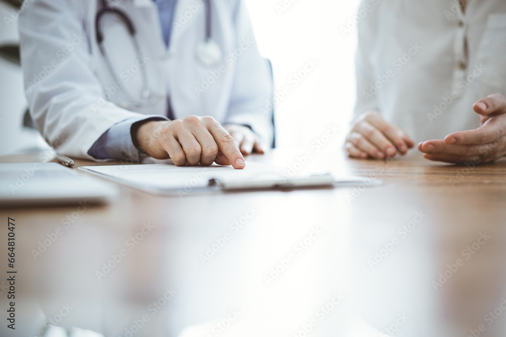 Doctor and patient sitting near each other at the wooden desk in clinic. Female physician's pointing to a records form. Medicine concept Doctor and patient sitting near each other at the wooden desk in clinic. Female physician's pointing to a records form. Medicine concept
