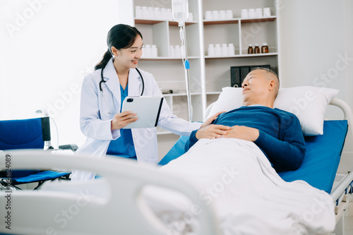 Asian doctor in white suit take notes while discussing and Asian elderly  man patient who lying on bed with receiving saline solution in hospital .