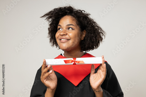 Smiling curly woman in graduate gown with paper in hands on white background