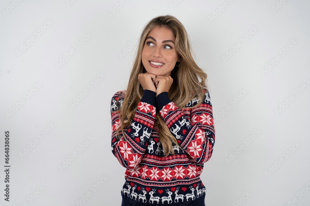 Happy Beautiful blonde girl wearing christmas sweater anticipates something awesome happen, looks happily aside, keeps hands together near face, has glad expression. Happy Beautiful blonde girl wearing christmas sweater anticipates something awesome happen, looks happily aside, keeps hands together near face, has glad expression.
