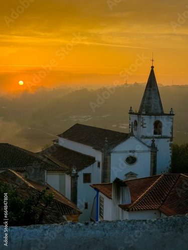 a sunset over a large white church surrounded by mountains and trees