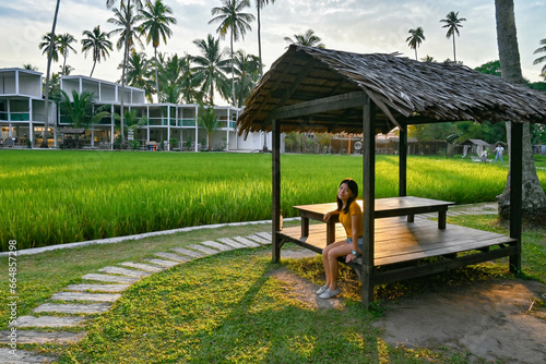 A girl sits in the gazebo beside the green paddy field in the evening.