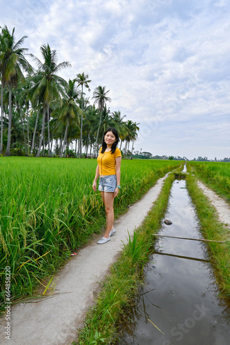 A girl stands beside the green paddy field and a small stream.