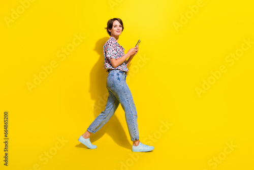 Full size profile photo of nice positive girl use smart phone walking empty space isolated on yellow color background