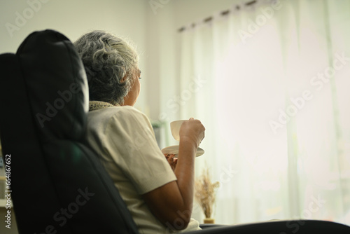Calm and peaceful elderly woman with cup of coffee sitting on armchair and looking away