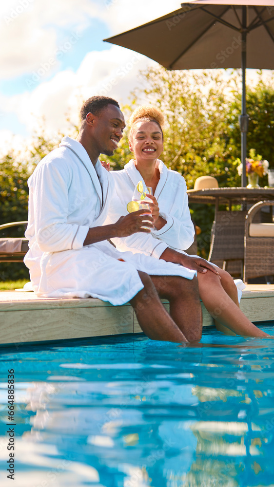 Couple Wearing Robes Outdoors Sitting With Drinks With Feet In Swimming Pool On Spa Day