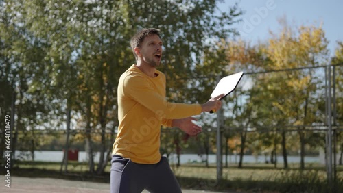 Dynamic shot of pickleball game on city court in park, handheld shot. Man actively receives passes and hits white ball with wooden racket. Maintaining active and healthy lifestyle.
