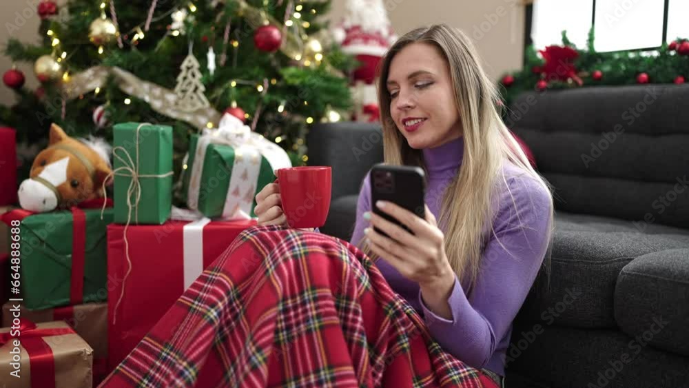 Young blonde woman using smartphone drinking coffee sitting by christmas tree at home Young blonde woman using smartphone drinking coffee sitting by christmas tree at home