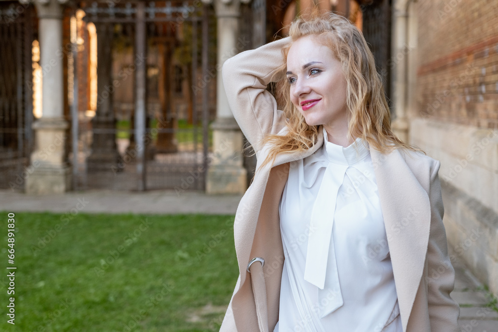 Fototapeta premium Portrait of attractive red-haired girl in spring outdoors. Cute blonde girl in white blouse and coat outside smiling and looking at the camera