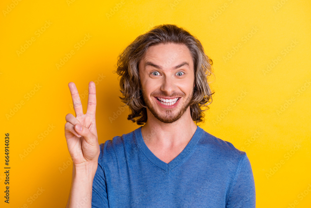 Photo of positive good mood man wear blue sweater smiling showing v-sign isolated yellow color background Photo of positive good mood man wear blue sweater smiling showing v-sign isolated yellow color background