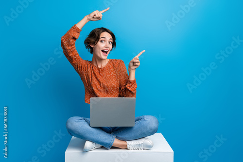 Full length photo of lovely young lady sit cube point empty space netbook wear trendy brown garment isolated on blue color background