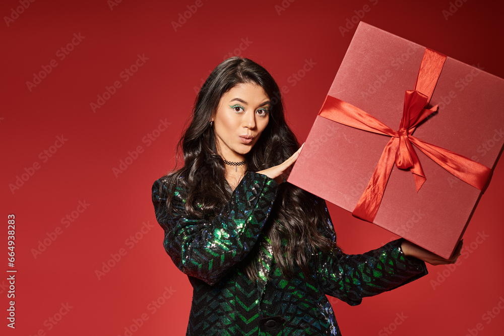 surprised asian woman in green jacket with sequins holding gift box on red backdrop, Merry Christmas surprised asian woman in green jacket with sequins holding gift box on red backdrop, Merry Christmas