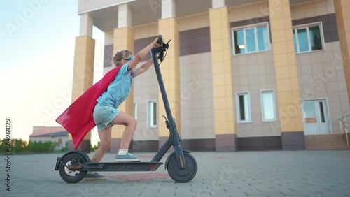 Girl superhero on electric scooter. happy family kid concept. a child on electric scooter rides on a large square in the city center under a blue sky with clouds and rays of sun. superhero girl dream