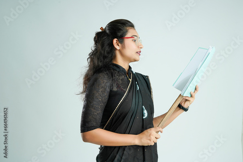Portrait of Indian lady teacher in saree stands against white or blackboard , conducting online class using Camera, internet and lights
