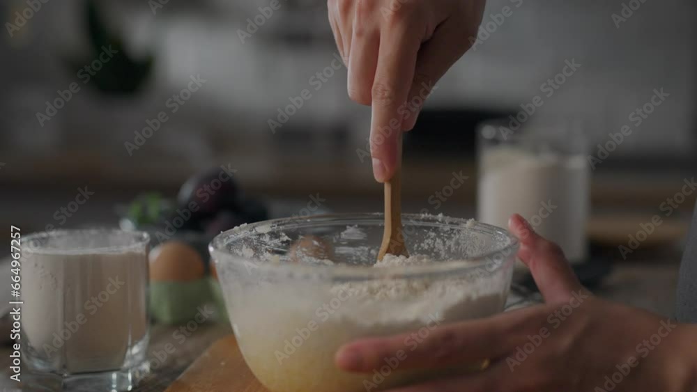 Woman is kneading dough in glass bowl while preparing dough for plum pie. Process of making fruit pie. Hands using wooden spoon to mix flour, knead dough. Sweet baking during plum season Woman is kneading dough in glass bowl while preparing dough for plum pie. Process of making fruit pie. Hands using wooden spoon to mix flour, knead dough. Sweet baking during plum season