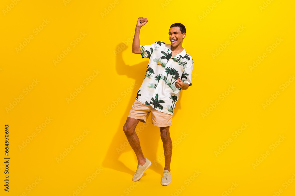 Full body photo of attractive young man raise fists celebration wear trendy palms print clothes isolated on yellow color background Full body photo of attractive young man raise fists celebration wear trendy palms print clothes isolated on yellow color background