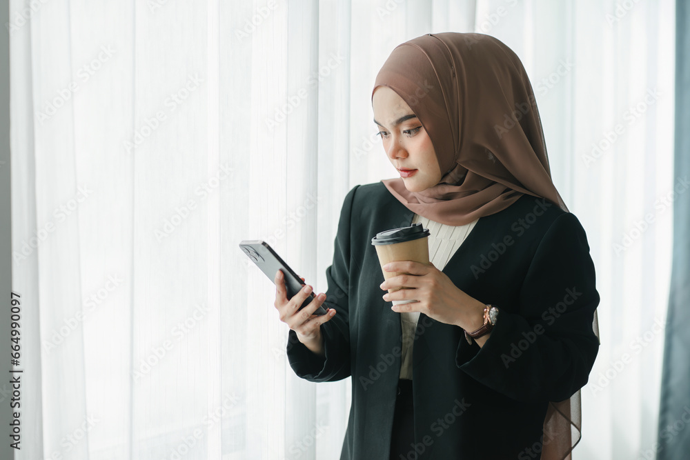 Asian Muslim businesswoman using a smartphone and enjoying coffee while standing in a modern office. Asian Muslim businesswoman using a smartphone and enjoying coffee while standing in a modern office.