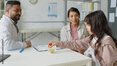 Waist up of Middle Eastern male child practitioner giving candies to Latin little girl and telling treatment recommendations to her mam while having checkup appointment in modern office