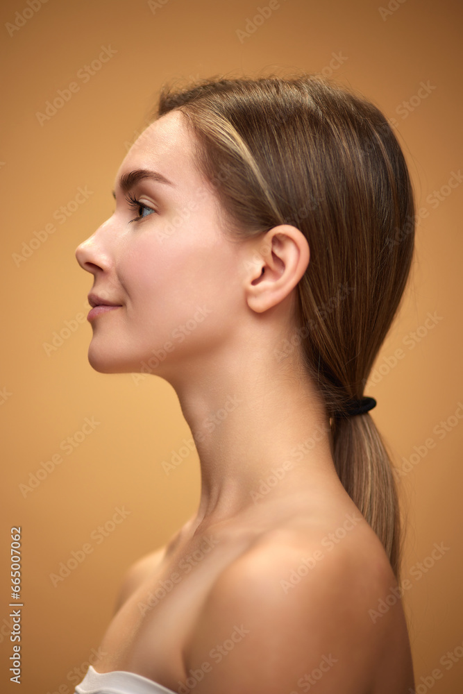 Profile view of tender, attractive young girl with ponytail, standing with bare shoulders isolated over light brown background. Concept of natural beauty, skincare, cosmetics and cosmetology Profile view of tender, attractive young girl with ponytail, standing with bare shoulders isolated over light brown background. Concept of natural beauty, skincare, cosmetics and cosmetology