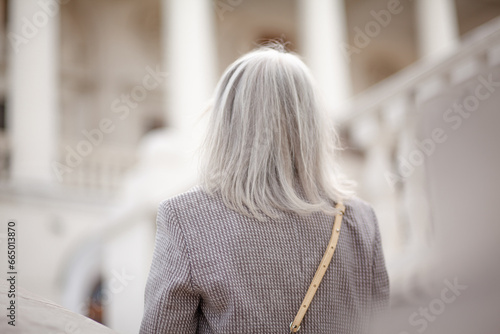 Back view of a  beauttiful woman with grey hair is walking arounfd the old town. A stylidh woman is wearing a gray coat, a skirt and a bag, photo