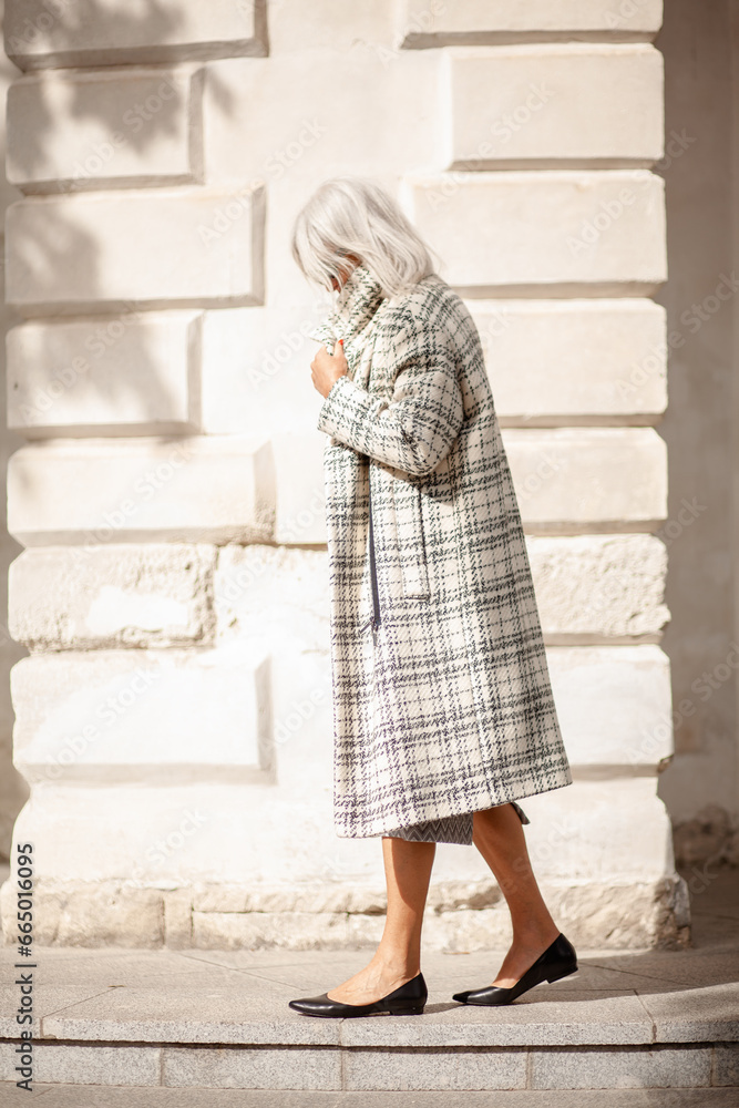 A  beauttiful woman with grey hair is walking arounв the old town. A stylidh woman is wearing a gray coat, a skirt and a bag and sunglasses