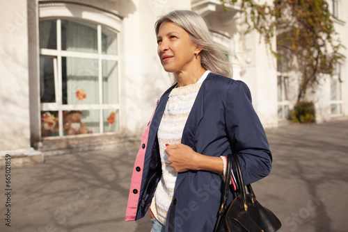 A  beauttiful woman with grey hair is walking around the old town. A stylidh woman is wearing a jeans, blue jacket and a bag