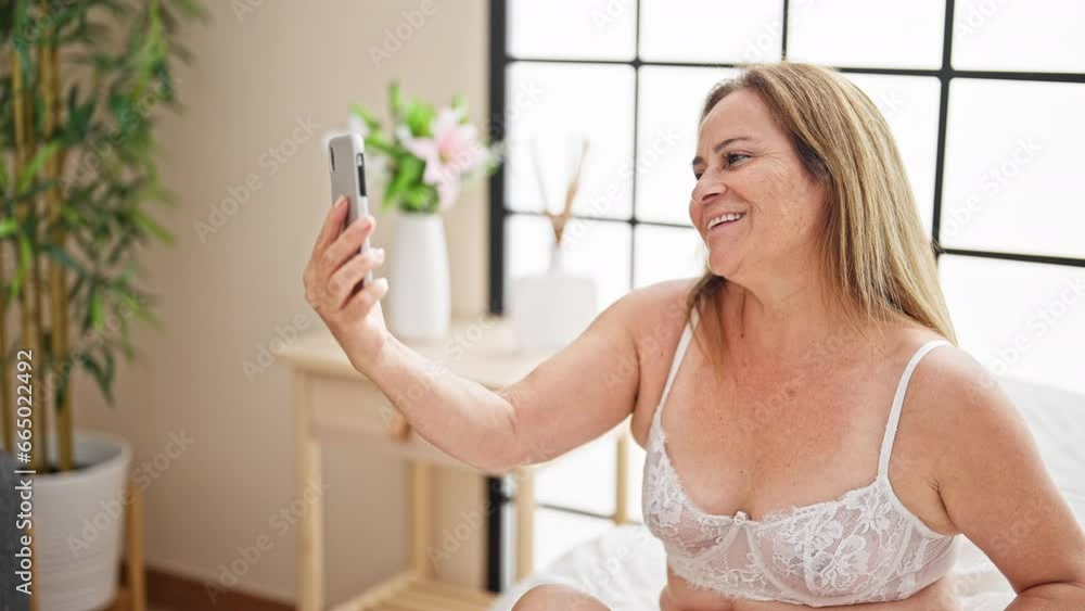 Middle age hispanic woman wearing sensual lingerie having video call sitting on bed at bedroom