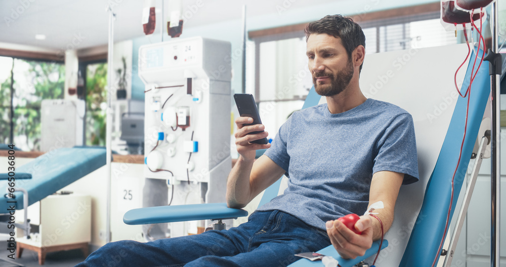Fototapeta premium Caucasian Man Donating Blood For People In Need In Hospital. Male Donor Squeezing Heart-Shaped Red Ball To Pump Blood And Chatting Online Using Smartphone. Donation For Organ Transplant Patients.