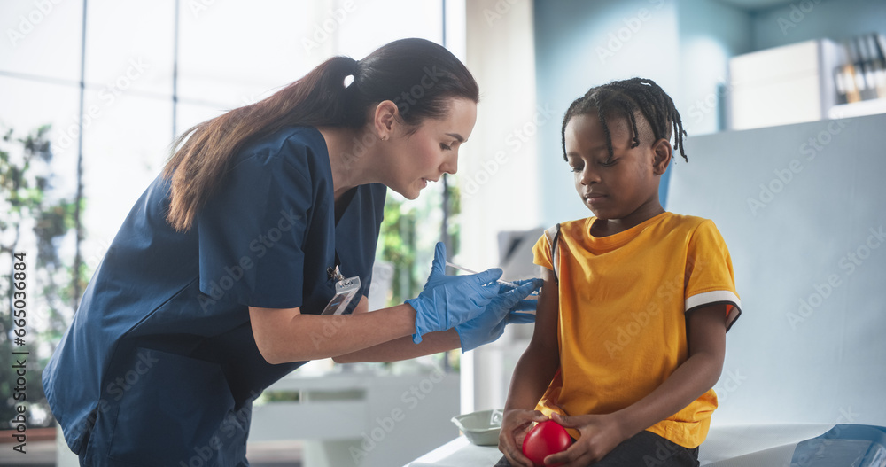 Obraz premium Young African American Boy Sitting In The Chair In Bright Hospital And Getting Polio Vaccine. Caucasian Female Nurse Is Performing Injection. Professional Woman Talks To Worried Kid, Calms Him Down.