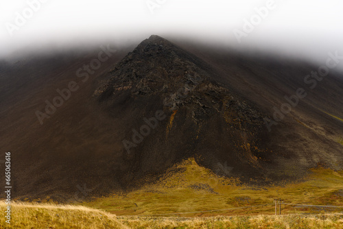 Rocky black volcanic mountain with peak visible through low-altitude clouds after heavy rain in Snaefellsnes peninsula in Iceland.